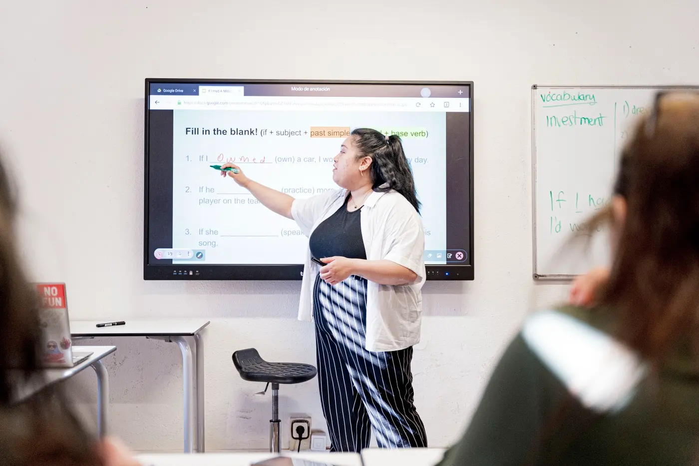 A trainee teacher pointing to a fill-in-the-blank grammar exercise on an interactive screen in front of her class